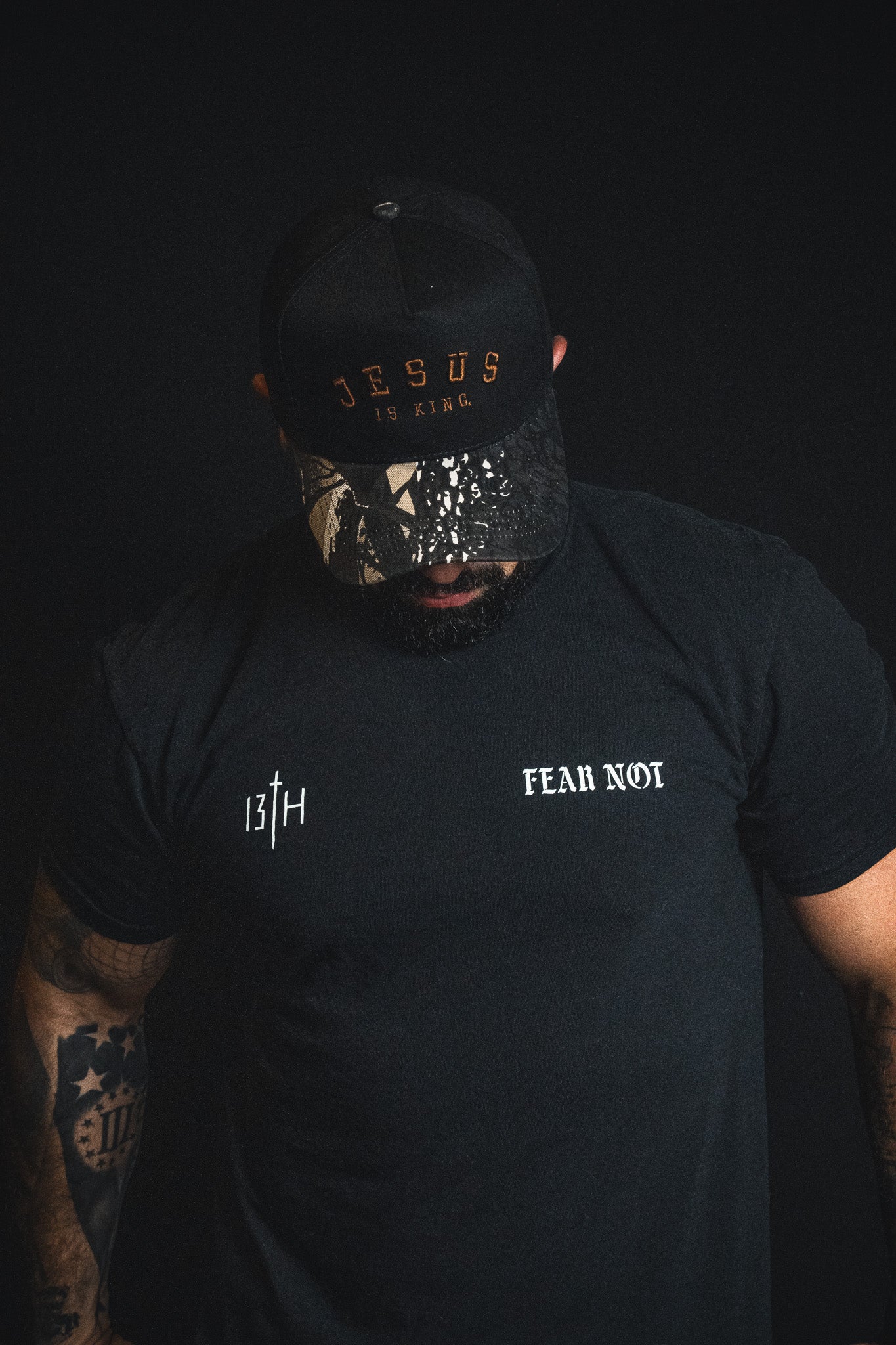 A man wearing a Born United Private Label “Jesus is King - Black and Darkwood Camo Snapback” and a black t-shirt with the words 13TH and FEAR NOT stands against a dark background, looking down.