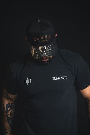 A man wearing a Born United Private Label “Jesus is King - Black and Darkwood Camo Snapback” and a black t-shirt with the words 13TH and FEAR NOT stands against a dark background, looking down.