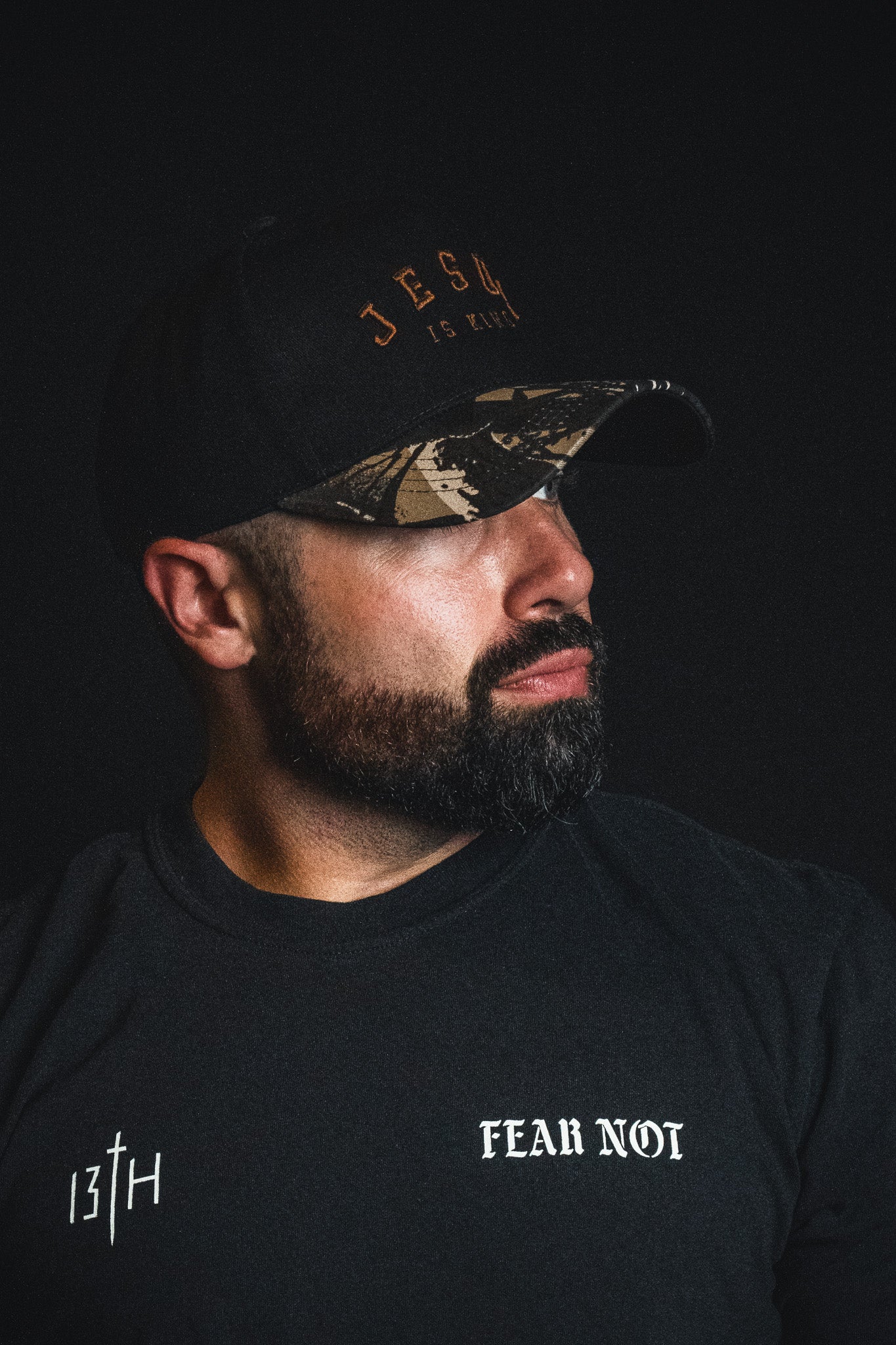 A man with a beard wearing a Born United Private Label Jesus is King - Black and Darkwood Camo Snapback and a black shirt that reads “13th” and “Fear Not,” set against a dark background.