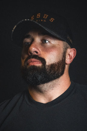 A man with a dark beard, dressed in a black shirt and wearing the Born United Private Label “Jesus is King - Black and Darkwood Camo Snapback,” gazes slightly to the side against a dark background.