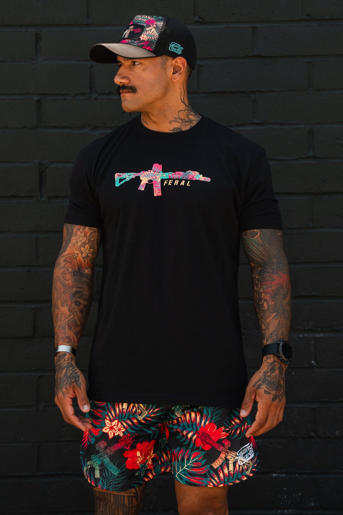 A tattooed man wears the Feral - Tee by Tango Charlie Apparel, featuring a colorful rifle graphic, paired with floral shorts and a matching cap, standing against a black brick wall and looking to his left.