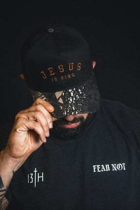 A man wears a Born United Private Label “Jesus is King - Black and Darkwood Camo Snapback” and a black shirt with “13TH” and “Fear Not” text, tilting his cap down against a dark background.