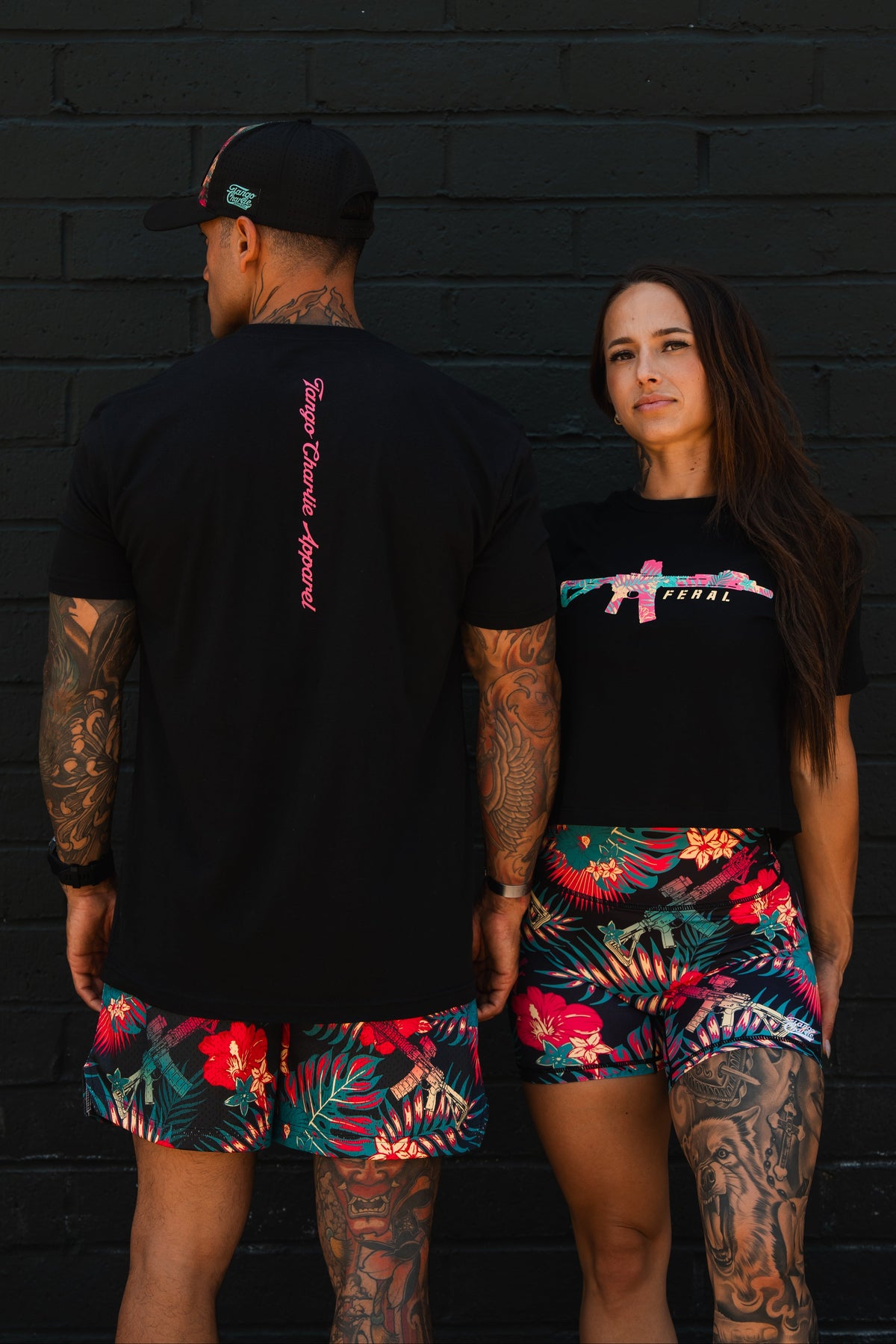 A man and woman pose by a black brick wall in matching vibrant floral shorts and black shirts. The woman stands confidently in the “Feral” Women’s Crop by Tango Charlie Apparel, celebrating bold, unfiltered women’s fashion.