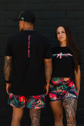 A man and woman pose by a black brick wall in matching vibrant floral shorts and black shirts. The woman stands confidently in the “Feral” Women’s Crop by Tango Charlie Apparel, celebrating bold, unfiltered women’s fashion.