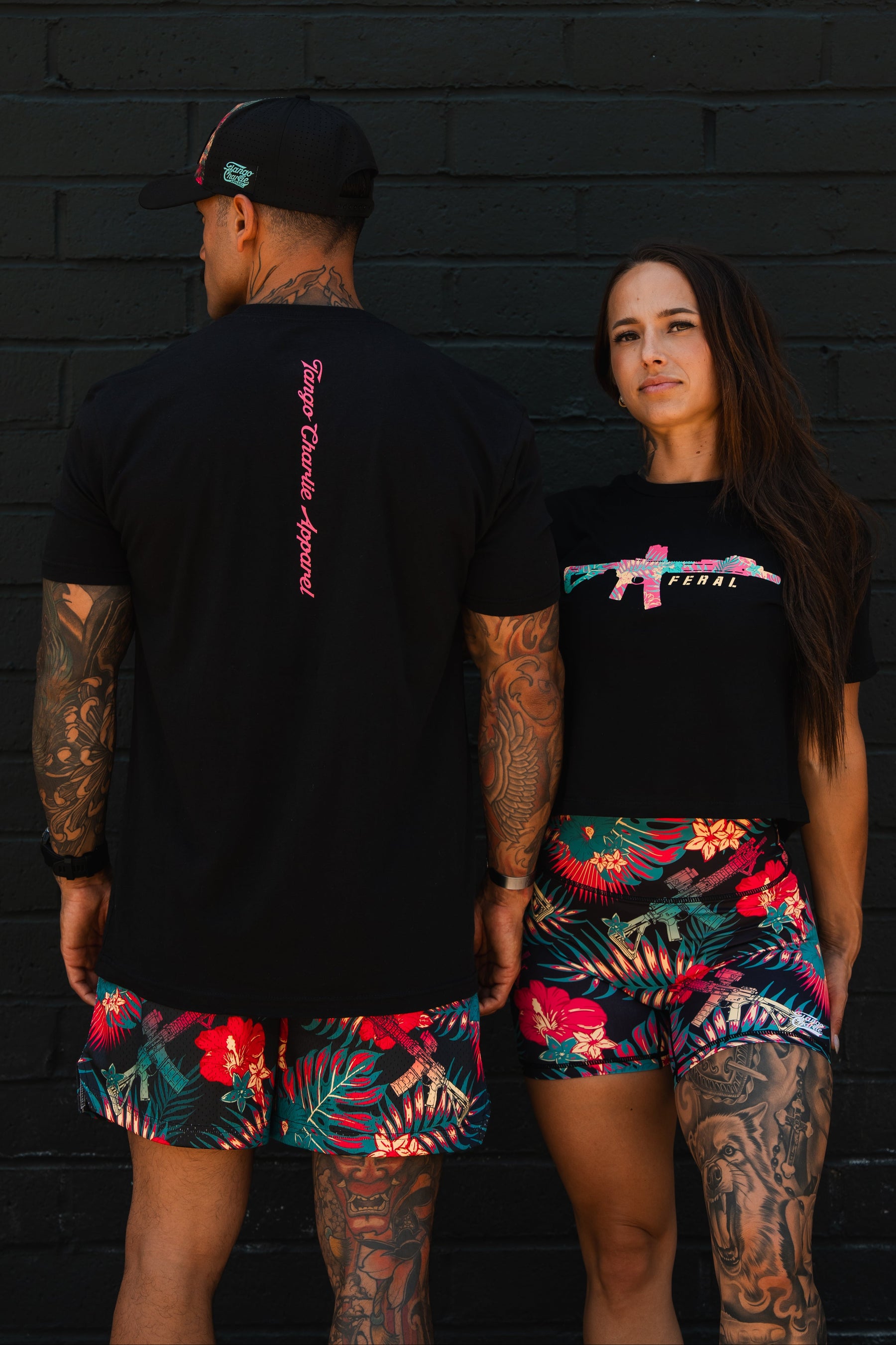 A man and woman in tropical floral shorts and black shirts with pink designs stand against a black brick wall. The woman wears the Feral - Women's Crop by Tango Charlie Apparel, confidently displaying her bold tattoos.
