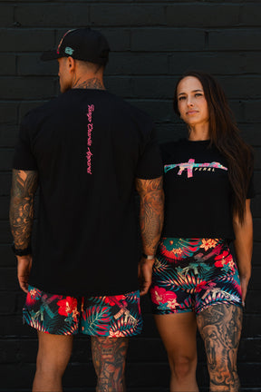 A man and woman in tropical floral shorts and black shirts with pink designs stand against a black brick wall. The woman wears the Feral - Women's Crop by Tango Charlie Apparel, confidently displaying her bold tattoos.