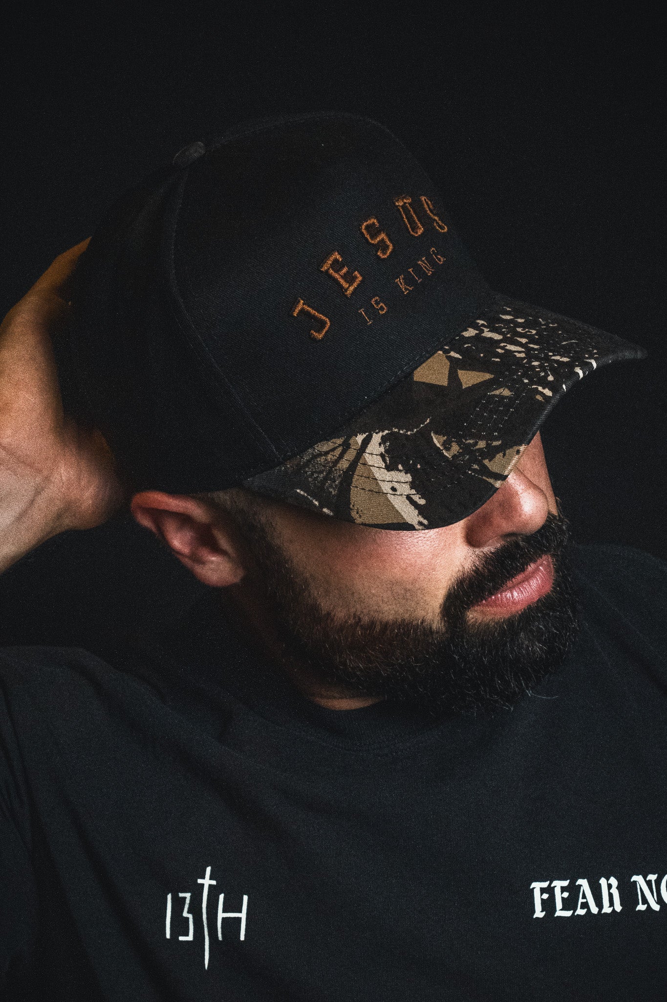 A man with a beard wears an embroidered Christian hat—a Born United Private Label “Jesus is King - Black and Darkwood Camo Snapback”—and a black shirt with “13TH” and “FEAR NO” text, set against a dark background.