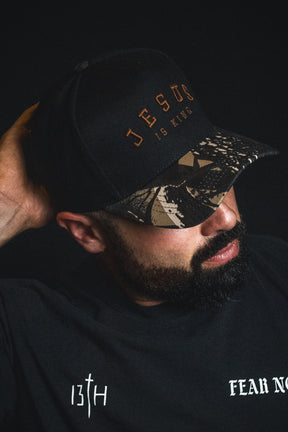 A man with a beard wears an embroidered Christian hat—a Born United Private Label “Jesus is King - Black and Darkwood Camo Snapback”—and a black shirt with “13TH” and “FEAR NO” text, set against a dark background.