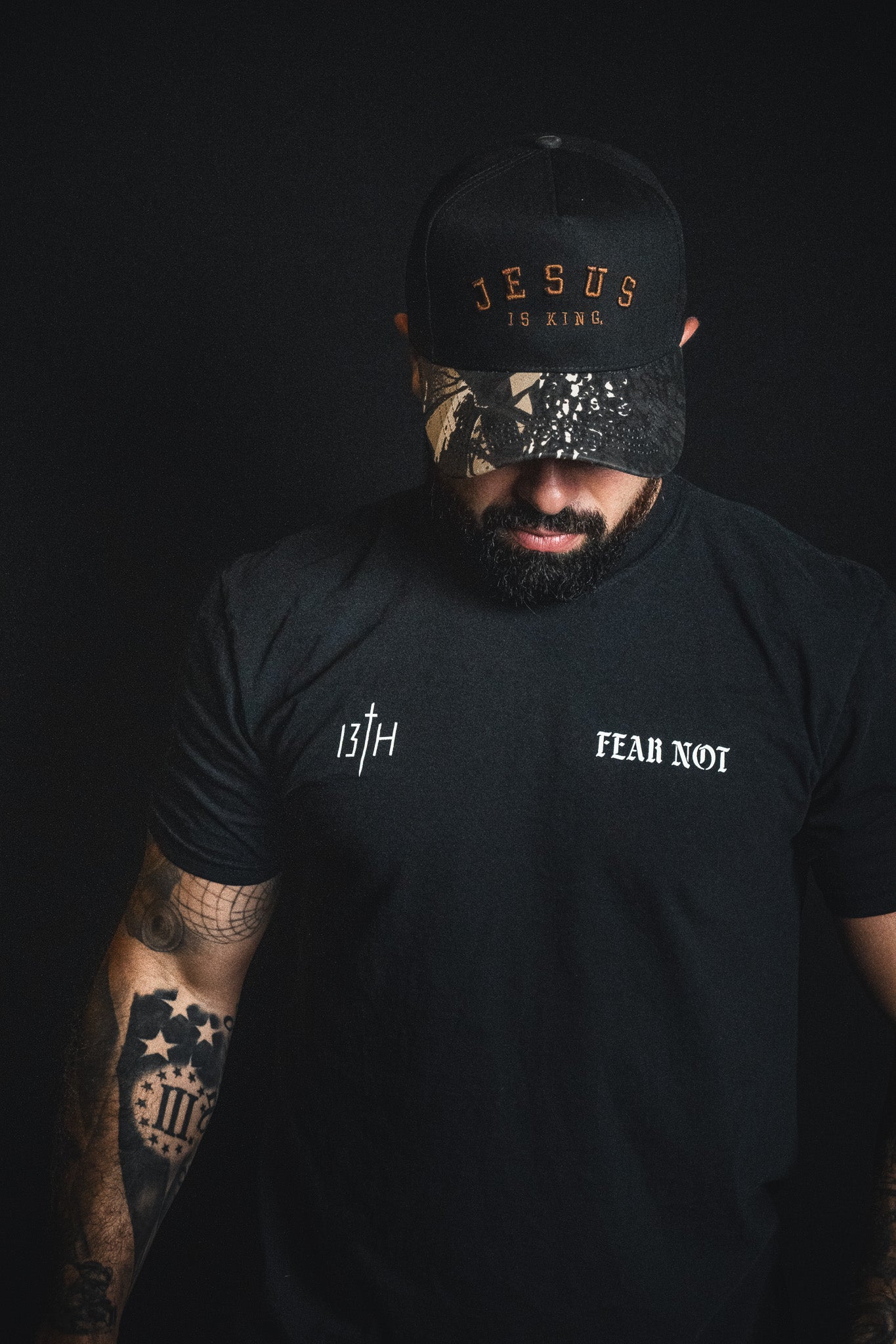 A man wearing a Born United Private Label Jesus is King - Black and Darkwood Camo Snapback and a black Fear Not t-shirt stands against a dark background, looking down.