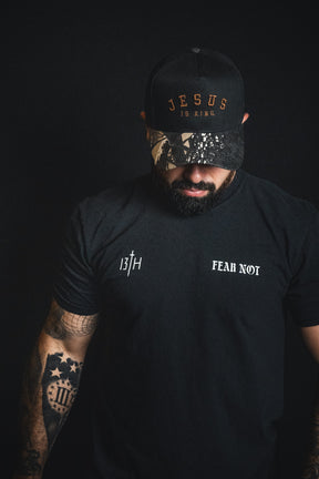 A man wearing a Born United Private Label Jesus is King - Black and Darkwood Camo Snapback and a black Fear Not t-shirt stands against a dark background, looking down.