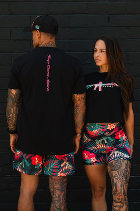 Two tattooed individuals in black shirts and vibrant tropical shorts stand before a black brick wall. The woman confidently faces forward, wearing the Feral - Women's Crop by Tango Charlie Apparel, showcasing bold women’s fashion.