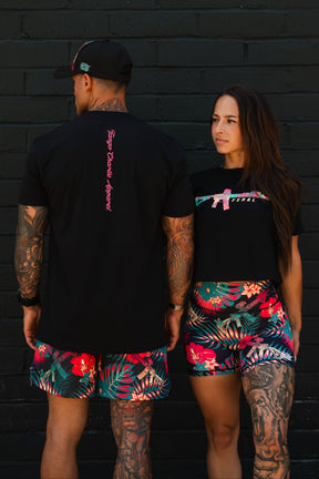 Two tattooed women in black shirts and vibrant tropical shorts stand against a dark brick wall. One wears the Feral - Women's Crop by Tango Charlie Apparel, both displaying bold patterns and unfiltered women's fashion with confidence.