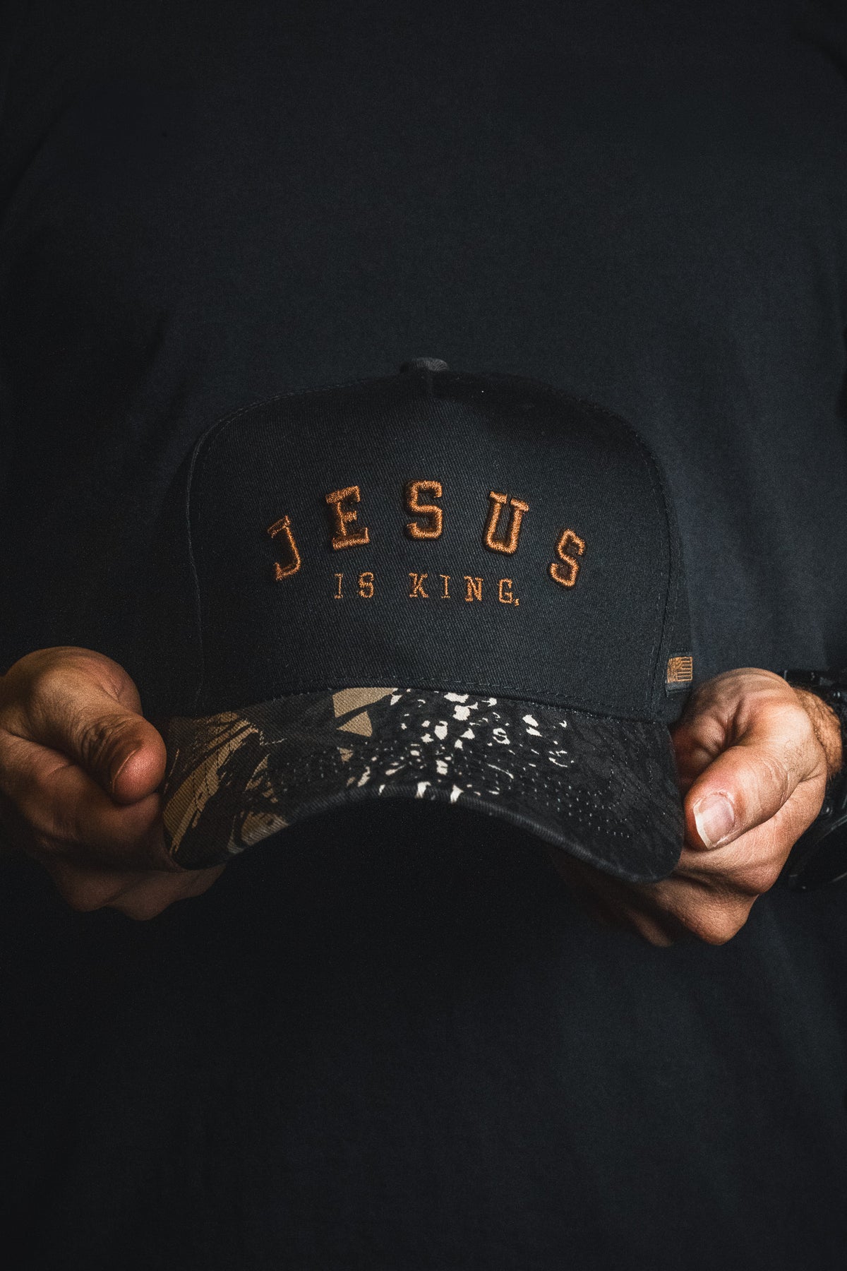 A person holds a Born United Private Label Jesus is King - Black and Darkwood Camo Snapback, featuring “JESUS IS KING” embroidered in orange letters and a darkwood camouflage-patterned brim.