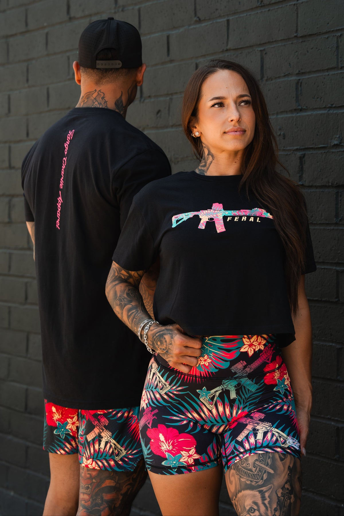 A woman in the Feral - Women's Crop by Tango Charlie Apparel stands by a dark brick wall, hand on hip, beside a man in bright floral shorts and a cap—showcasing bold, unfiltered women’s fashion.