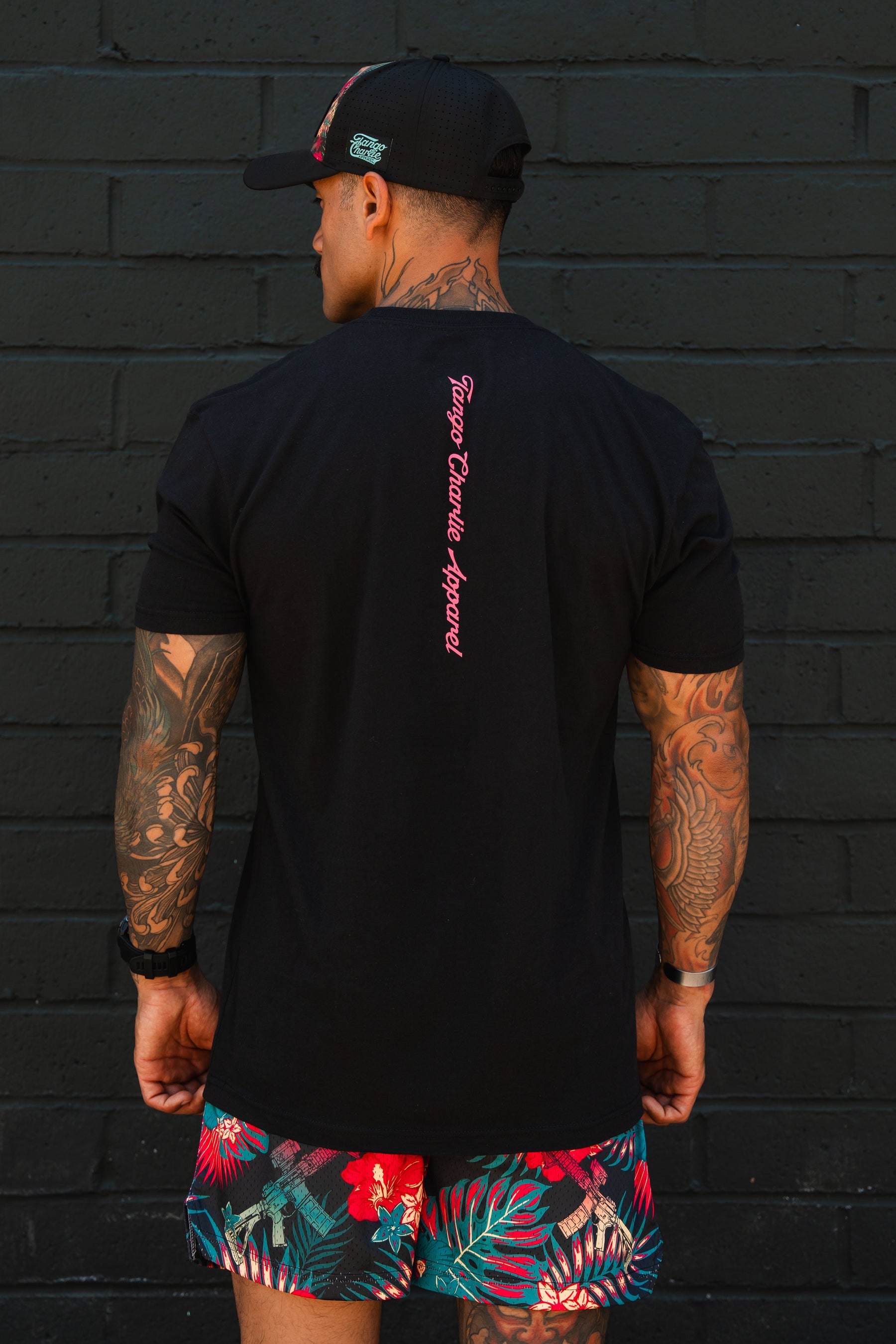 A man with tattooed arms stands before a black brick wall, wearing Tango Charlie Apparel's "Feral - Tee," a black T-shirt featuring pink text down the back, paired with a black cap and vibrant tropical shorts with red and green patterns.