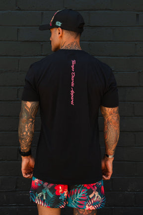 A man with tattooed arms stands before a black brick wall, wearing Tango Charlie Apparel's "Feral - Tee," a black T-shirt featuring pink text down the back, paired with a black cap and vibrant tropical shorts with red and green patterns.