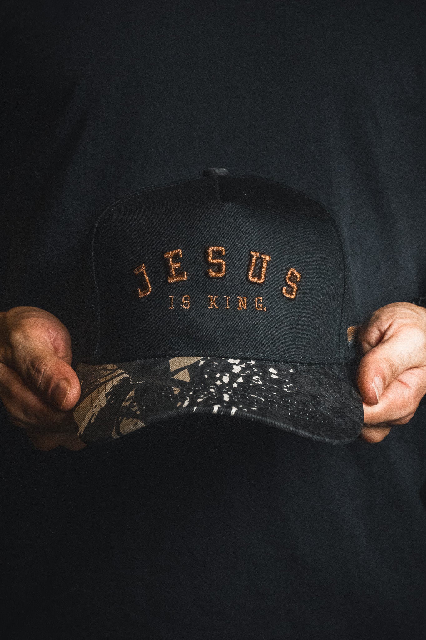 A person holds the Jesus is King - Black and Darkwood Camo Snapback by Born United Private Label—a black cap with brown “JESUS IS KING” lettering and a patterned brim—against a dark background.