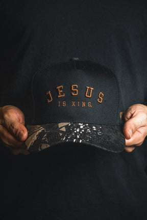 A person holds the Jesus is King - Black and Darkwood Camo Snapback by Born United Private Label—a black cap with brown “JESUS IS KING” lettering and a patterned brim—against a dark background.