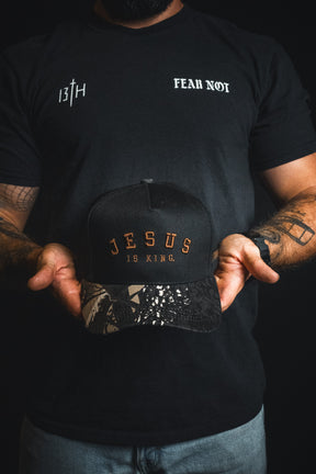 A person holds the Born United Private Label “Jesus is King - Black and Darkwood Camo Snapback,” an embroidered Christian hat, while wearing a black shirt with “FEAR NOT” and “13TH” printed on the chest.