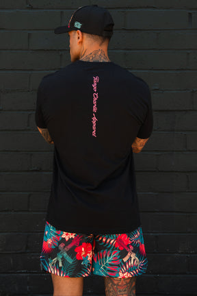 Wearing the Tango Charlie Apparel Feral - Tee, a black cap, and tropical print shorts, a person displays tattoos on their arms and neck against a dark brick wall—an image of radical authenticity.