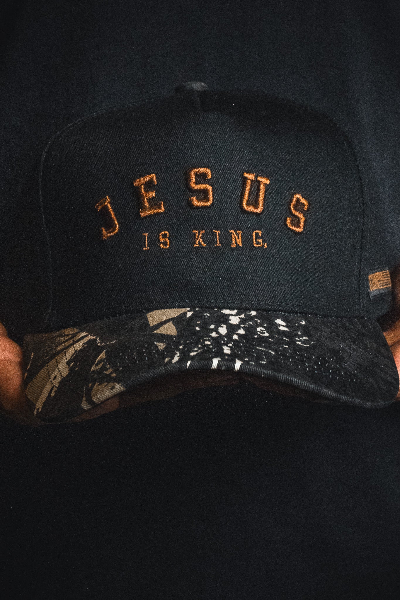 A Jesus is King - Black and Darkwood Camo Snapback by Born United Private Label, featuring “JESUS IS KING.” embroidered in orange on the front and a patterned camo baseball cap-style brim, held by someone against a dark background.