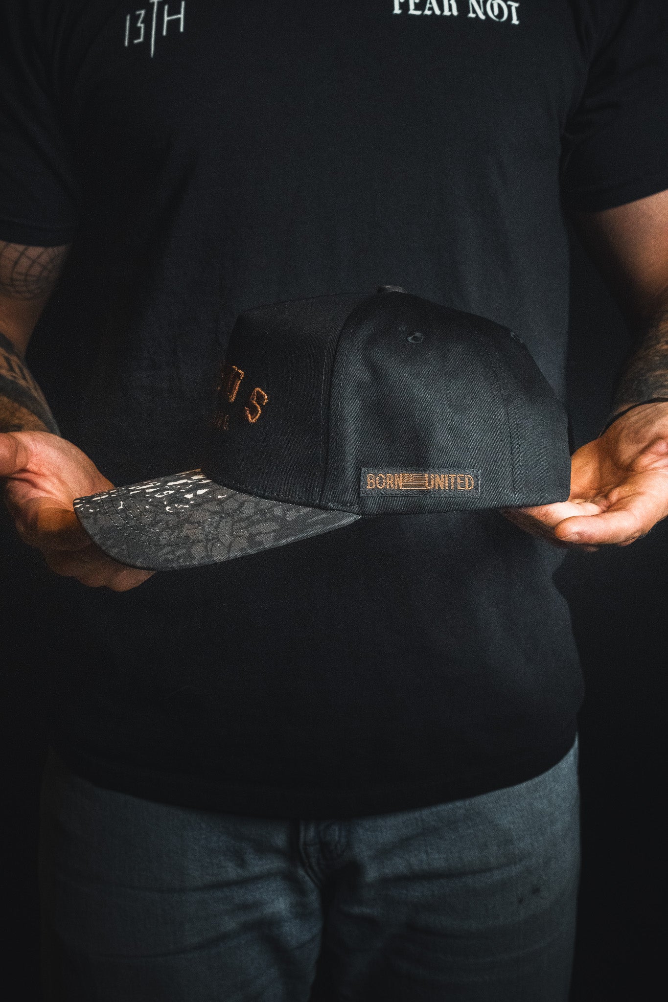 A person wearing a black shirt and jeans holds the Jesus is King - Black and Darkwood Camo Snapback by Born United Private Label in both hands against a dark background.