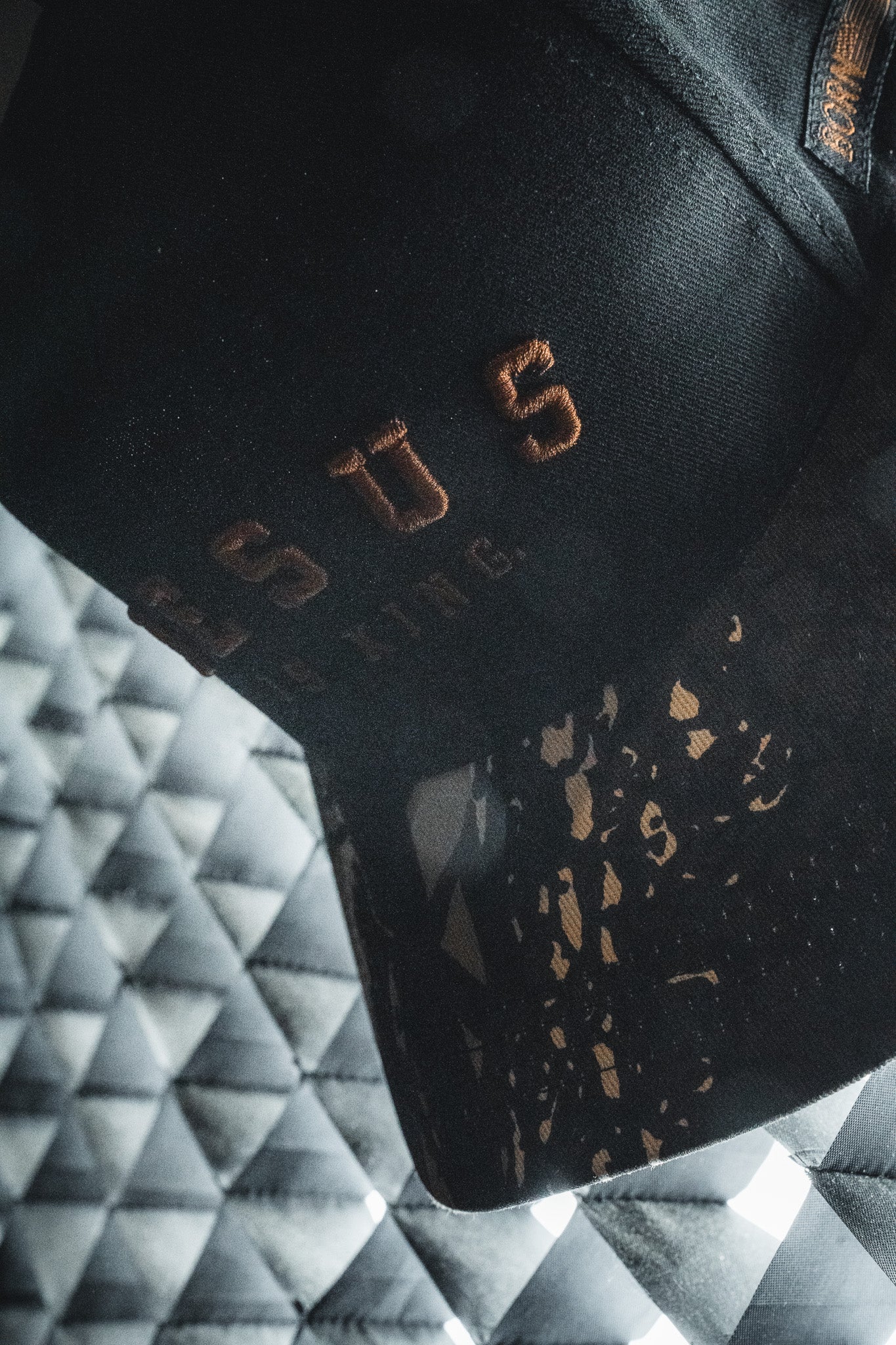 Certainly! Here’s a rewritten sentence using the provided product data: A Born United Private Label “Jesus is King” Black and Darkwood Camo Snapback rests on a textured, gray, quilted surface highlighted by patterns of light and shadow.