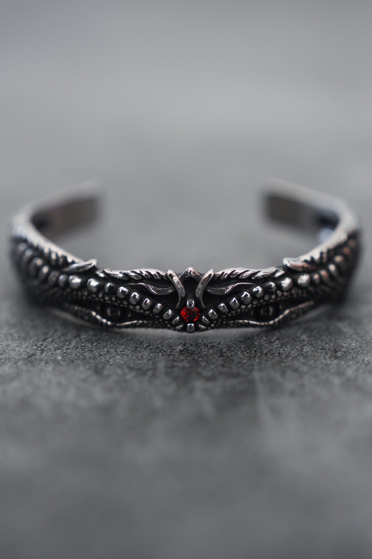 A close-up of the intricately designed Relentless Betrayal Holy Grail Bracelet showcases its dark stainless steel with a dragon motif, detailed scales, wings, and a central red gem. The softly blurred gray background enhances its mythical artifact allure.