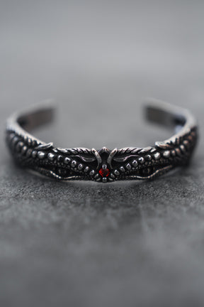 A close-up of the intricately designed Relentless Betrayal Holy Grail Bracelet showcases its dark stainless steel with a dragon motif, detailed scales, wings, and a central red gem. The softly blurred gray background enhances its mythical artifact allure.
