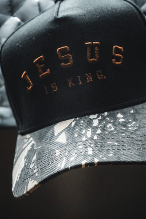 The Born United Private Label “Jesus is King - Black and Darkwood Camo Snapback” features “JESUS IS KING.” embroidered in gold on the front, with a brim displaying a black, white, and gray abstract Darkwood camo pattern.
