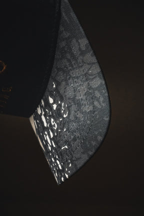 A close-up of the “Jesus is King - Black and Darkwood Camo Snapback” by Born United Private Label, a black baseball cap with a curved brim featuring a white speckled camo pattern, designed in the style of an embroidered Christian hat.