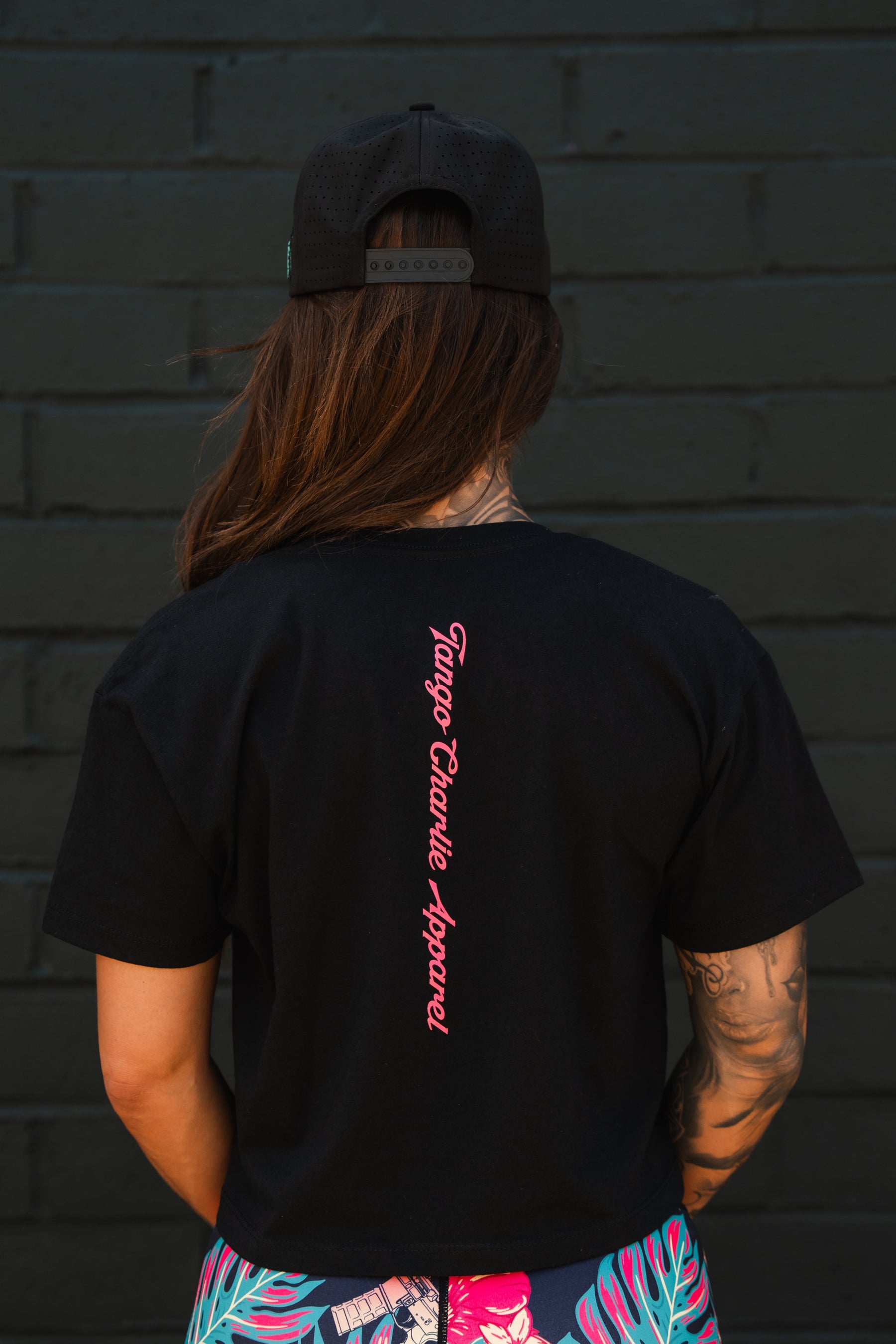 A woman with long brown hair, black cap, and an arm tattoo wears the Feral - Women's Crop by Tango Charlie Apparel, featuring pink branding on the back, as she stands before a dark brick wall—showcasing bold women’s fashion.