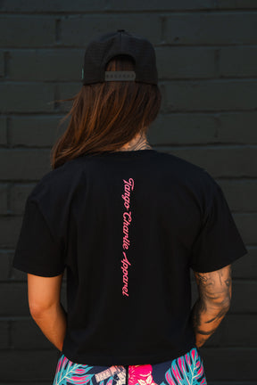 A woman with long brown hair, black cap, and an arm tattoo wears the Feral - Women's Crop by Tango Charlie Apparel, featuring pink branding on the back, as she stands before a dark brick wall—showcasing bold women’s fashion.