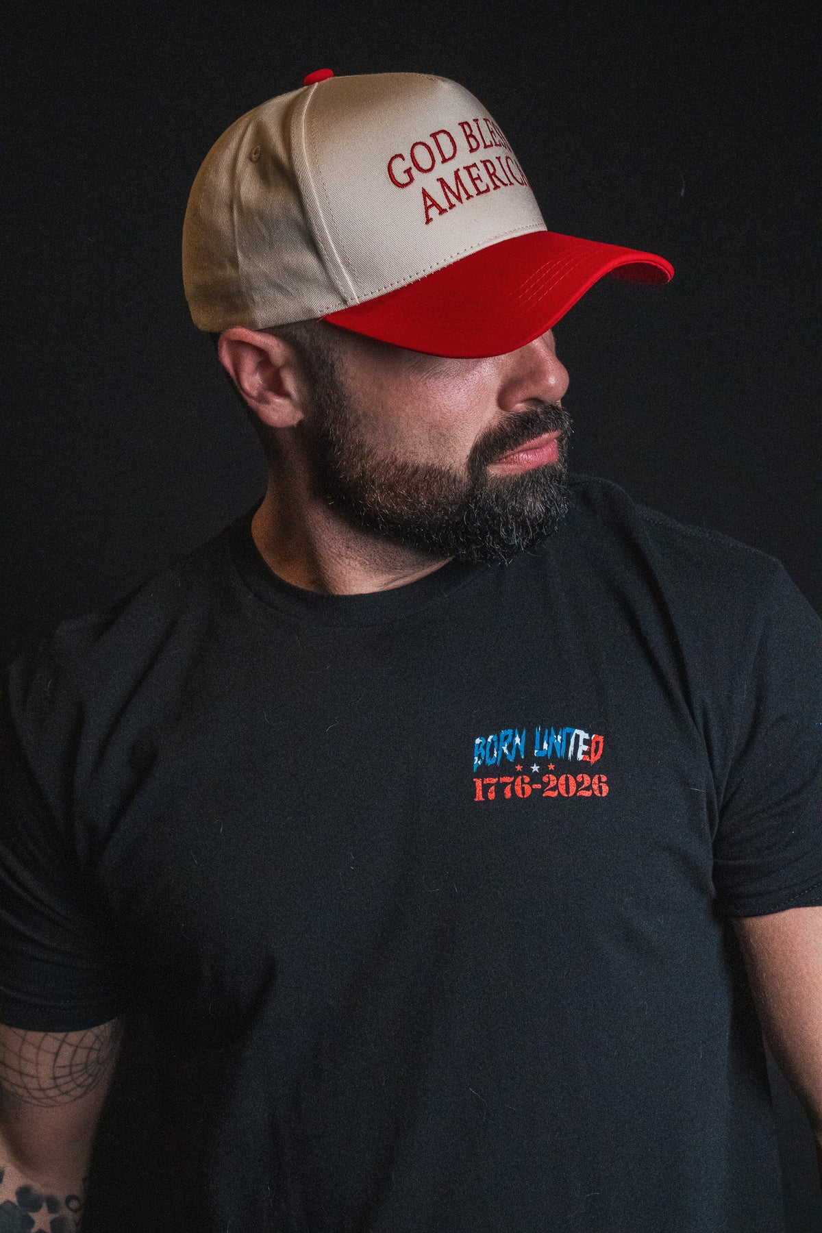 A man wearing a black Born United 1776-2026 t-shirt and a Born United Private Label God Bless America Snapback stands against a black background.