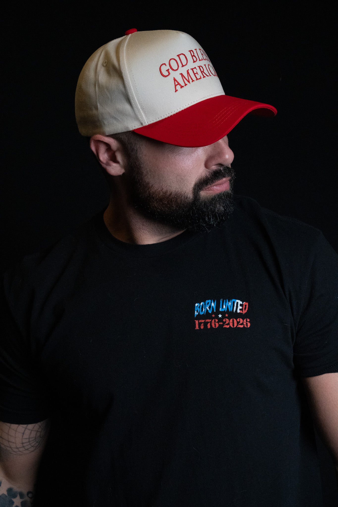 A bearded man wearing the Born United Private Label “God Bless America Snapback” in white and red and a black T-shirt with “BORN UNITED 1776-2026” printed on it, gazes to the side against a black background.