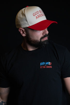 A bearded man wearing the Born United Private Label “God Bless America Snapback” in white and red and a black T-shirt with “BORN UNITED 1776-2026” printed on it, gazes to the side against a black background.