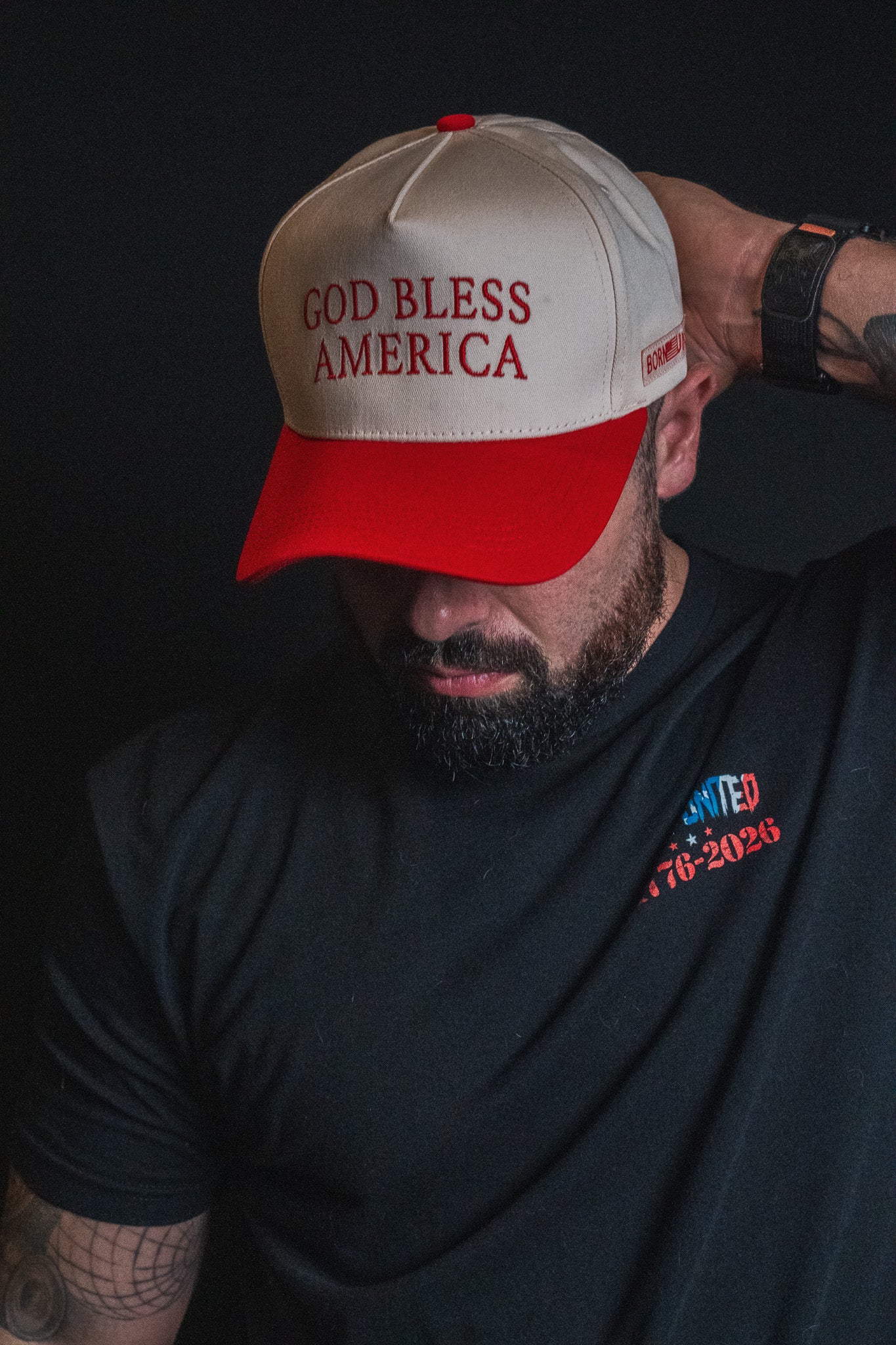 A man faces downward against a dark background, wearing the Born United Private Label God Bless America Snapback in red and white embroidery and a black shirt with the UNITED 1776-2026 logo.