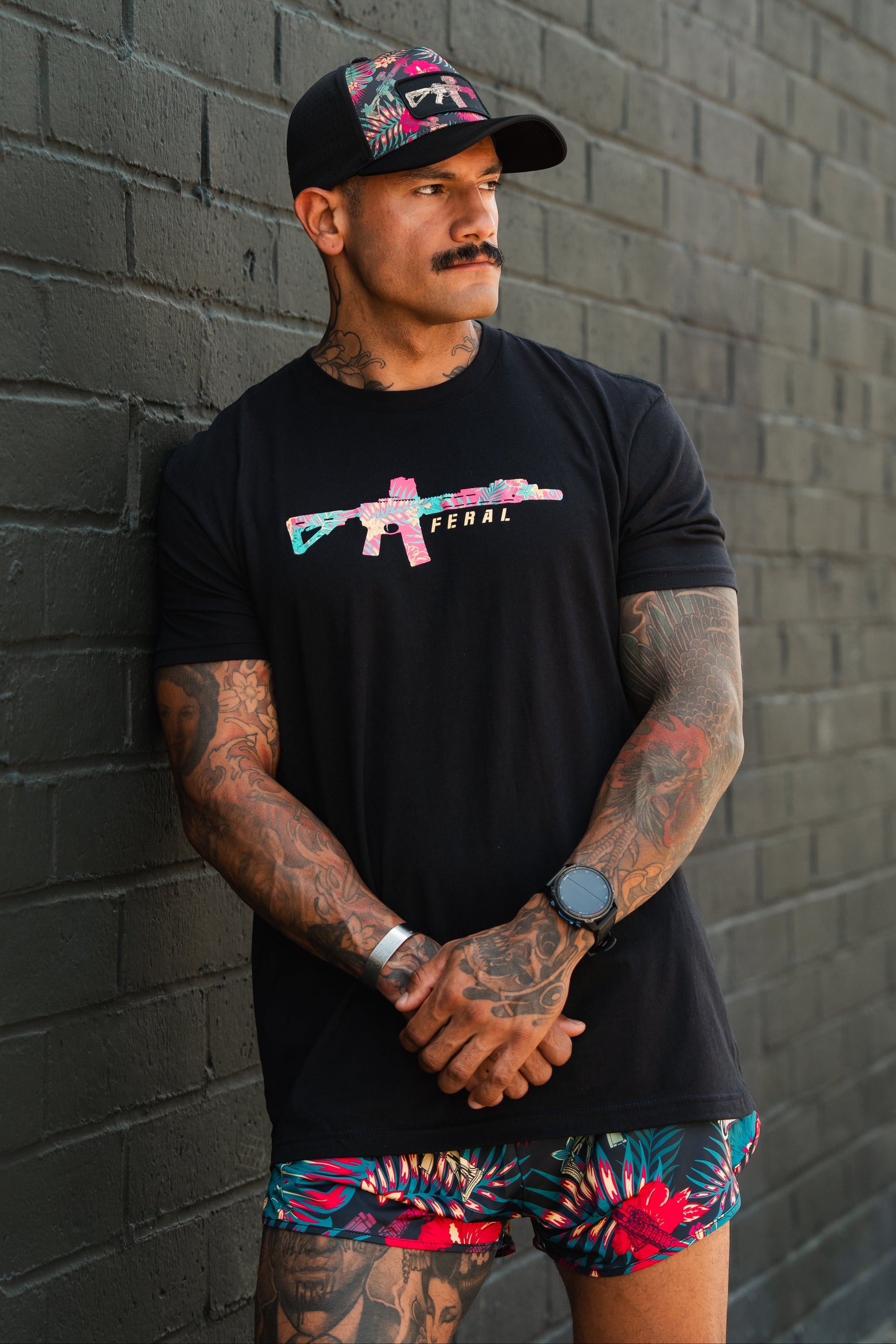 A tattooed man in a black cap stands against a brick wall, arms crossed. Wearing Tango Charlie Apparel's Feral - Tee with a vibrant gun graphic and floral shorts, he gazes to the side, embodying radical authenticity.