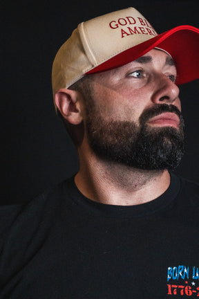 A man with a dark beard wears the Born United Private Label God Bless America Snapback in beige and red, paired with a black shirt, as he stands against a dark background looking off to the side.