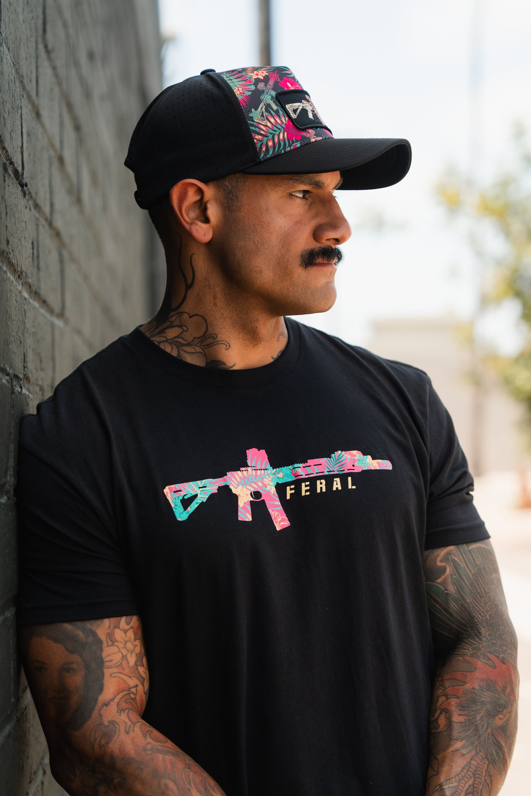 A tattooed, mustached man leans against a brick wall, wearing Tango Charlie Apparel's Feral - Tee with a vibrant rifle graphic and "FERAL" text, paired with a black floral cap for radical authenticity.