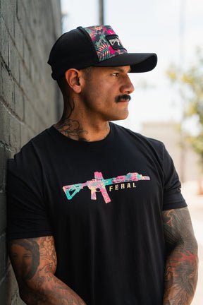 A tattooed, mustached man leans against a brick wall, wearing Tango Charlie Apparel's Feral - Tee with a vibrant rifle graphic and "FERAL" text, paired with a black floral cap for radical authenticity.