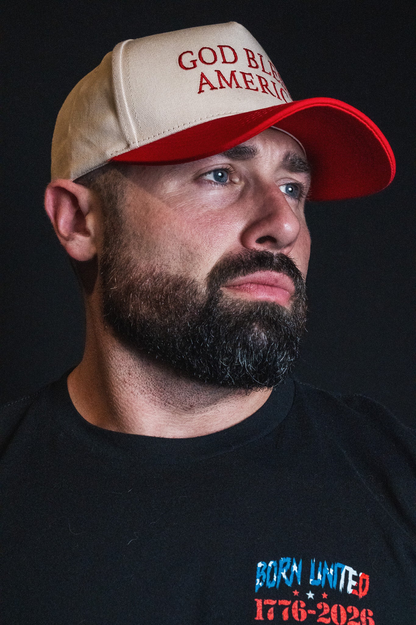 A bearded man, wearing the Born United Private Label God Bless America Snapback and a Born United 1776-2026 shirt, looks to the side against a dark background.