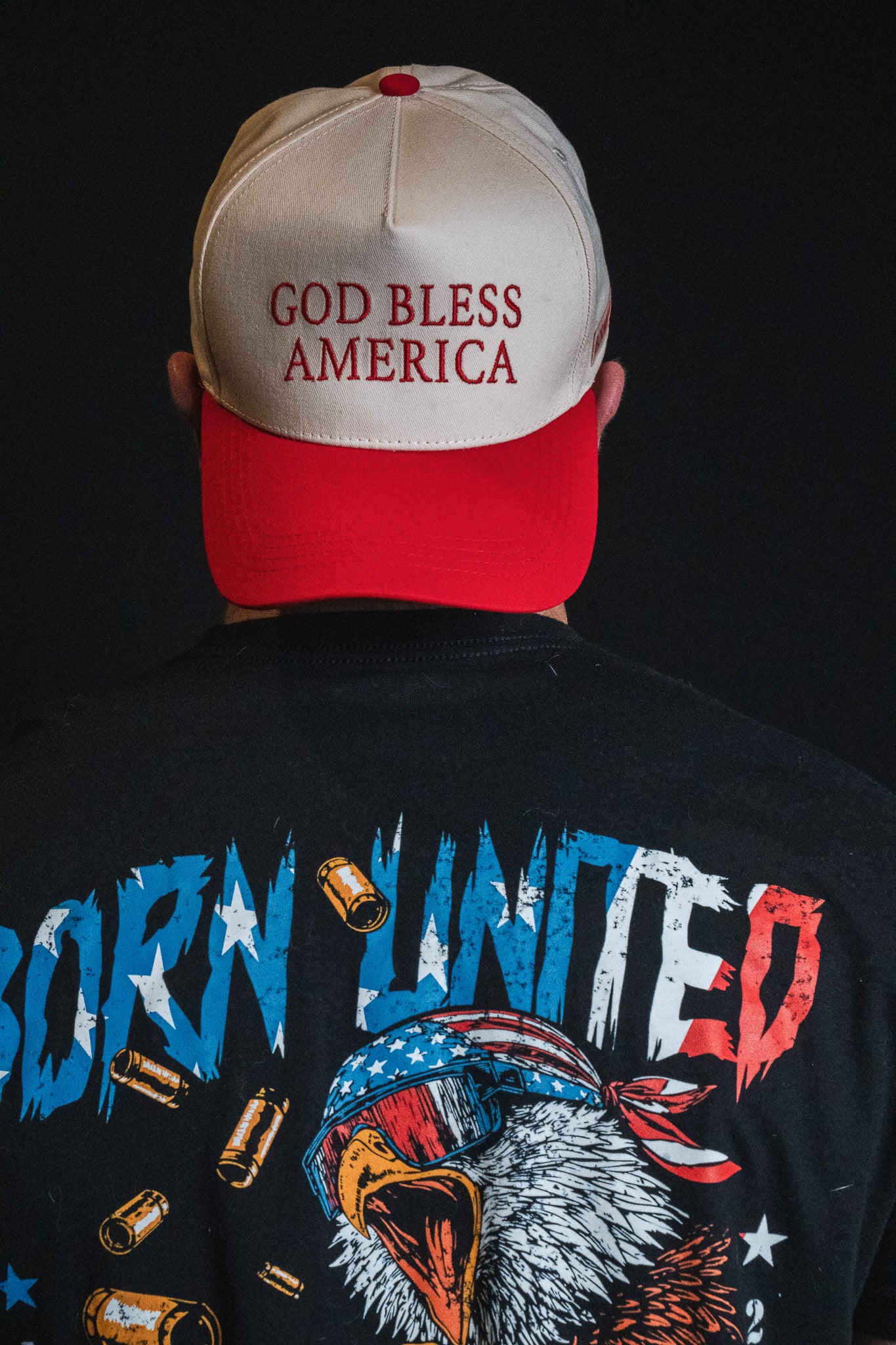 A person is facing away, wearing a God Bless America Snapback by Born United Private Label and a black shirt featuring an eagle, bullets, and the words “Born United.”.