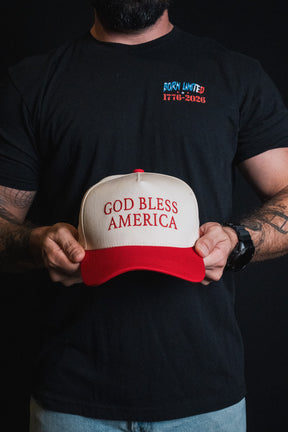 A tattooed individual holds a God Bless America Snapback by Born United Private Label, wearing a black BORN UNITED 1776-2026 T-shirt against a black background.