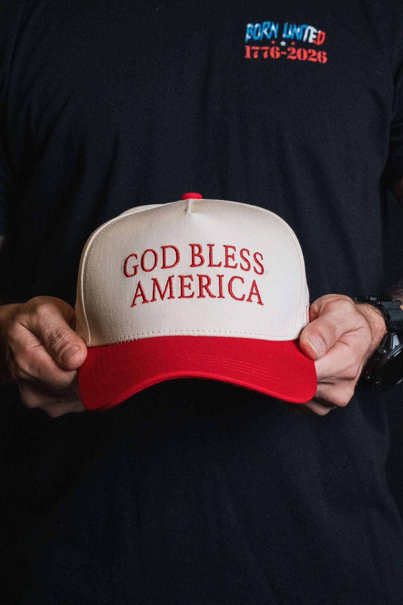 A person holding the white and red Born United Private Label God Bless America Snapback, featuring bold red embroidery, while wearing a black shirt displaying the “BORN UNITED 1776-2026” text.