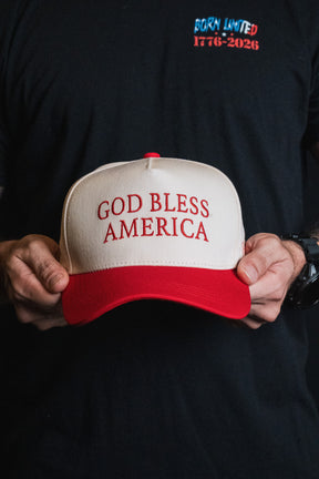 A person holding the white and red Born United Private Label God Bless America Snapback, featuring bold red embroidery, while wearing a black shirt displaying the “BORN UNITED 1776-2026” text.
