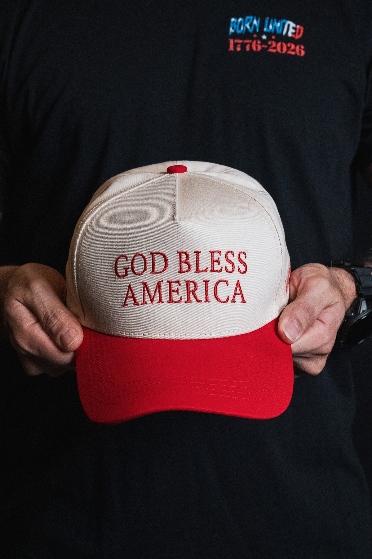 A person holds the Born United Private Label “God Bless America” snapback hat, which is white and red with embroidered text; they are wearing a black shirt featuring “BORN UNITED 1776–2026” in red and blue letters.