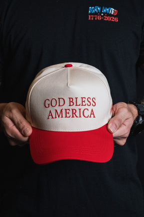 A person holds the Born United Private Label “God Bless America” snapback hat, which is white and red with embroidered text; they are wearing a black shirt featuring “BORN UNITED 1776–2026” in red and blue letters.