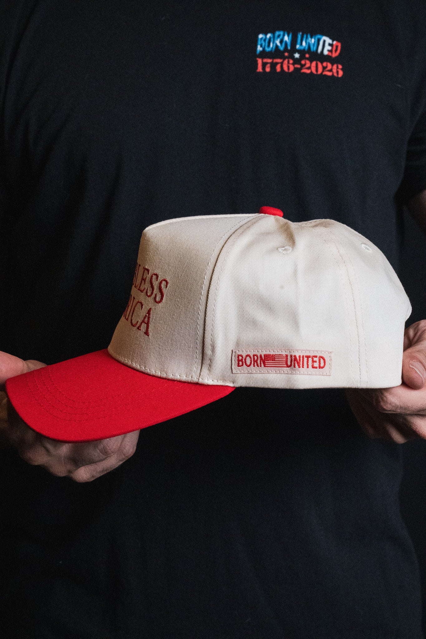 A person holds the God Bless America Snapback by Born United Private Label, a white and red baseball cap. They are wearing a black shirt with “BORN UNITED 1776-2026” printed in colorful text on the chest.