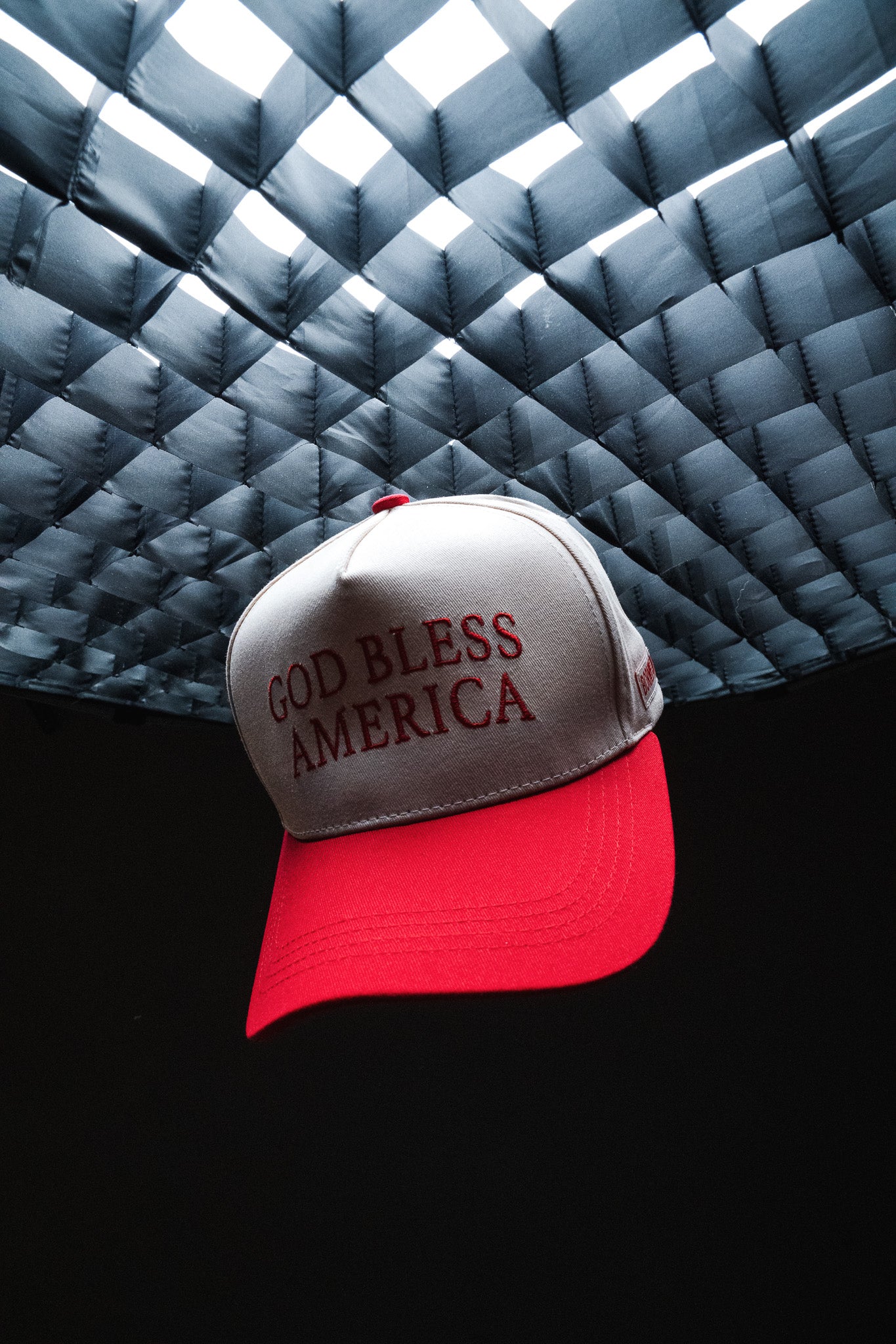 A red and white embroidered God Bless America Snapback by Born United Private Label floats under a woven, metallic-looking ceiling against a dark background, highlighting the bold text and classic style of this signature cap.